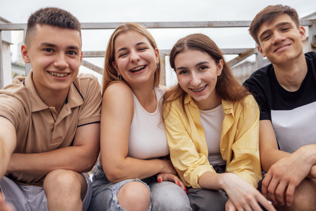 Four cute teenagers taking a selfie. Funny friends takes a picture of theyself on the phone outdoors. Smiling boys and girls sitting on the street. Happy teens enjoying vacation together.